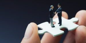 Business success strategy with collaboration, teamwork or negotiation jigsaw key, miniature people businessmen handshaking on white jigsaw puzzle piece in real human hand, dark black background.